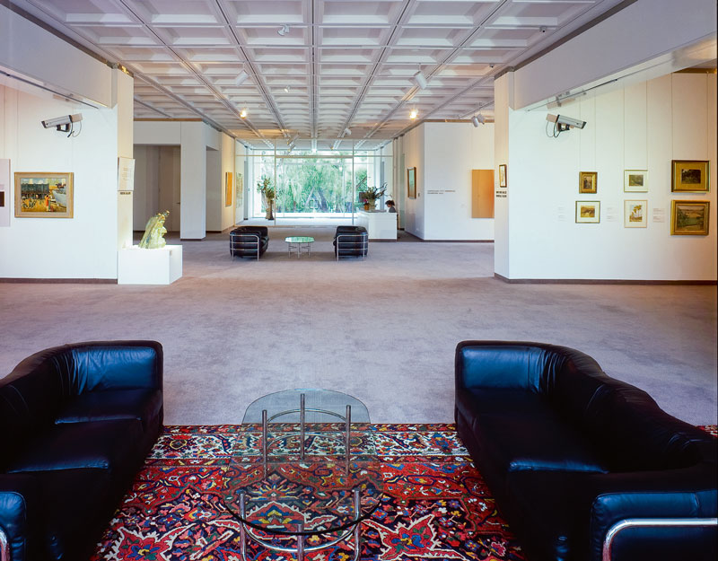 A photo of the interior of an art gallery. In the foreground are a pair of black leather couches, a glass coffee table and a patterned rug. In the distance you can see art hanging on the walls and the entrance to the gallery.