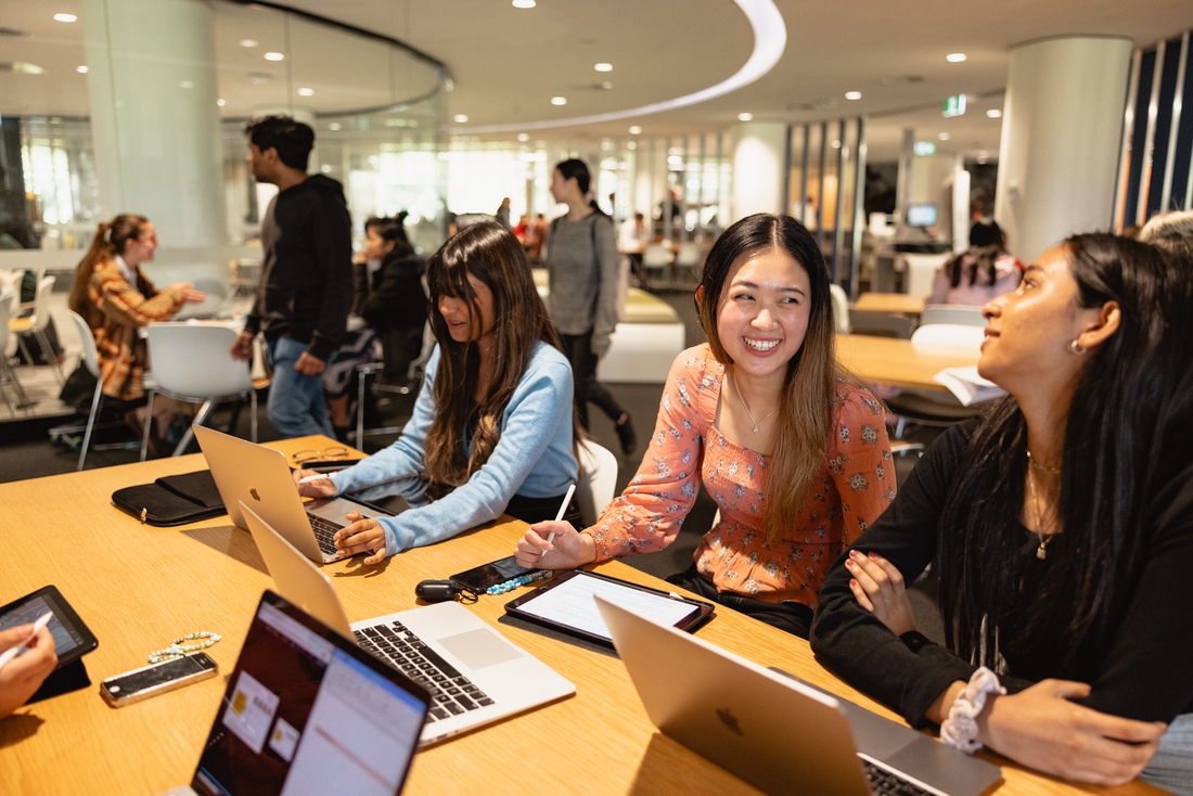 A group of students sitting at a desk with laptops in the Reid Library. The students are chatting and working together.