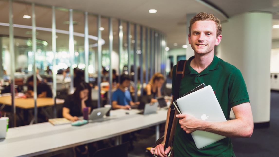 Student standing in library holding a laptop computer