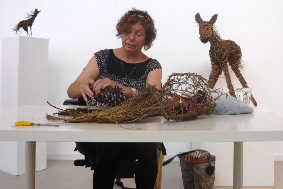 Photograph of a person seated at a table working on a woven sculpture with two finished woven animal sculptures behind them