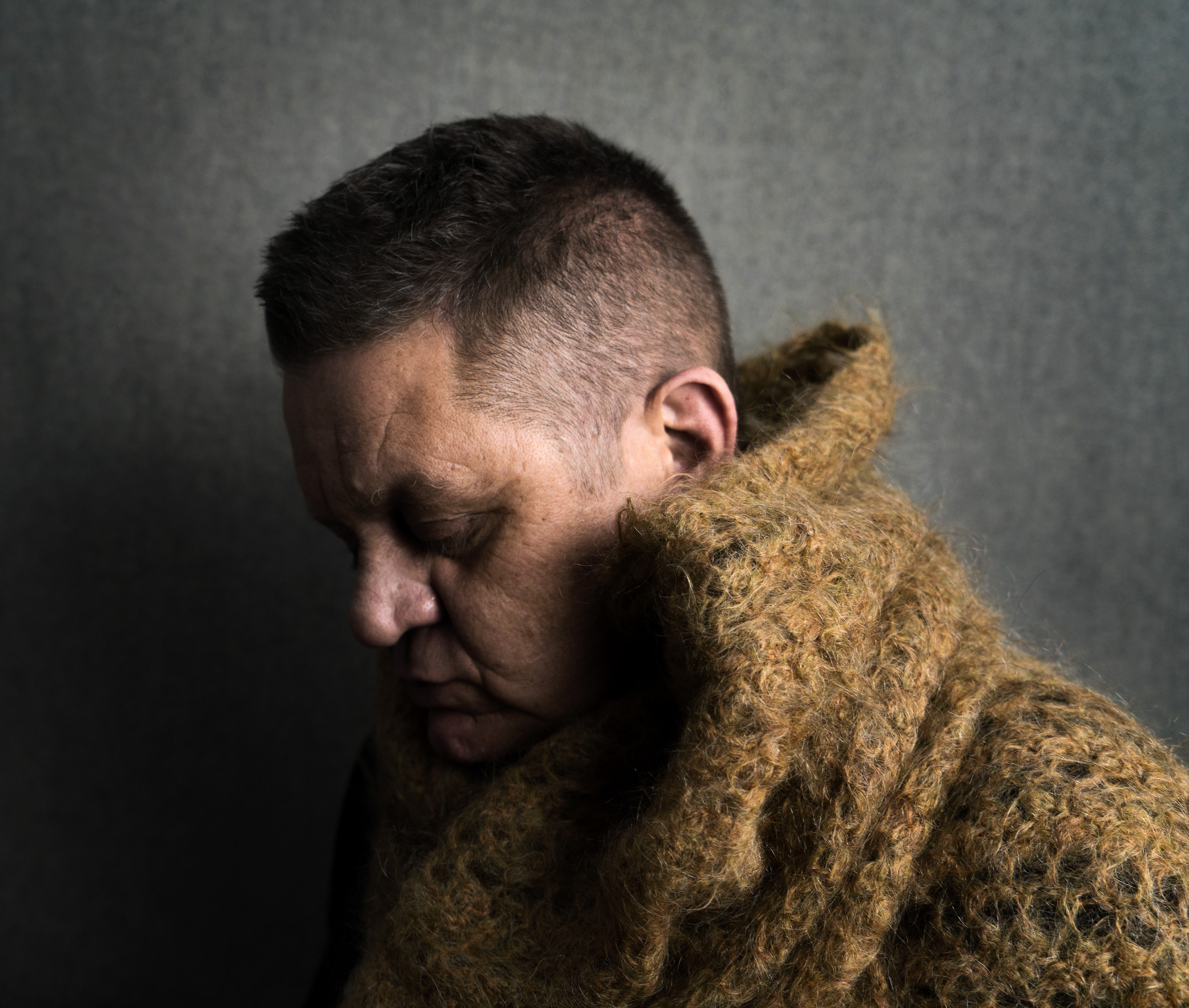 A person is photographed from the shoulders up in profile with eyes cast down and wearing a knit olive coat