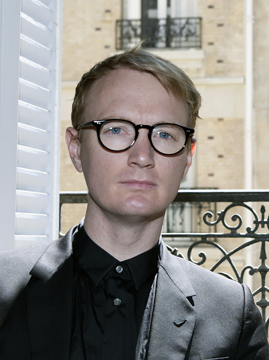 Photograph of a person from the shoulders up in a black collared shirt with black rimmed glasses looking at the camera 