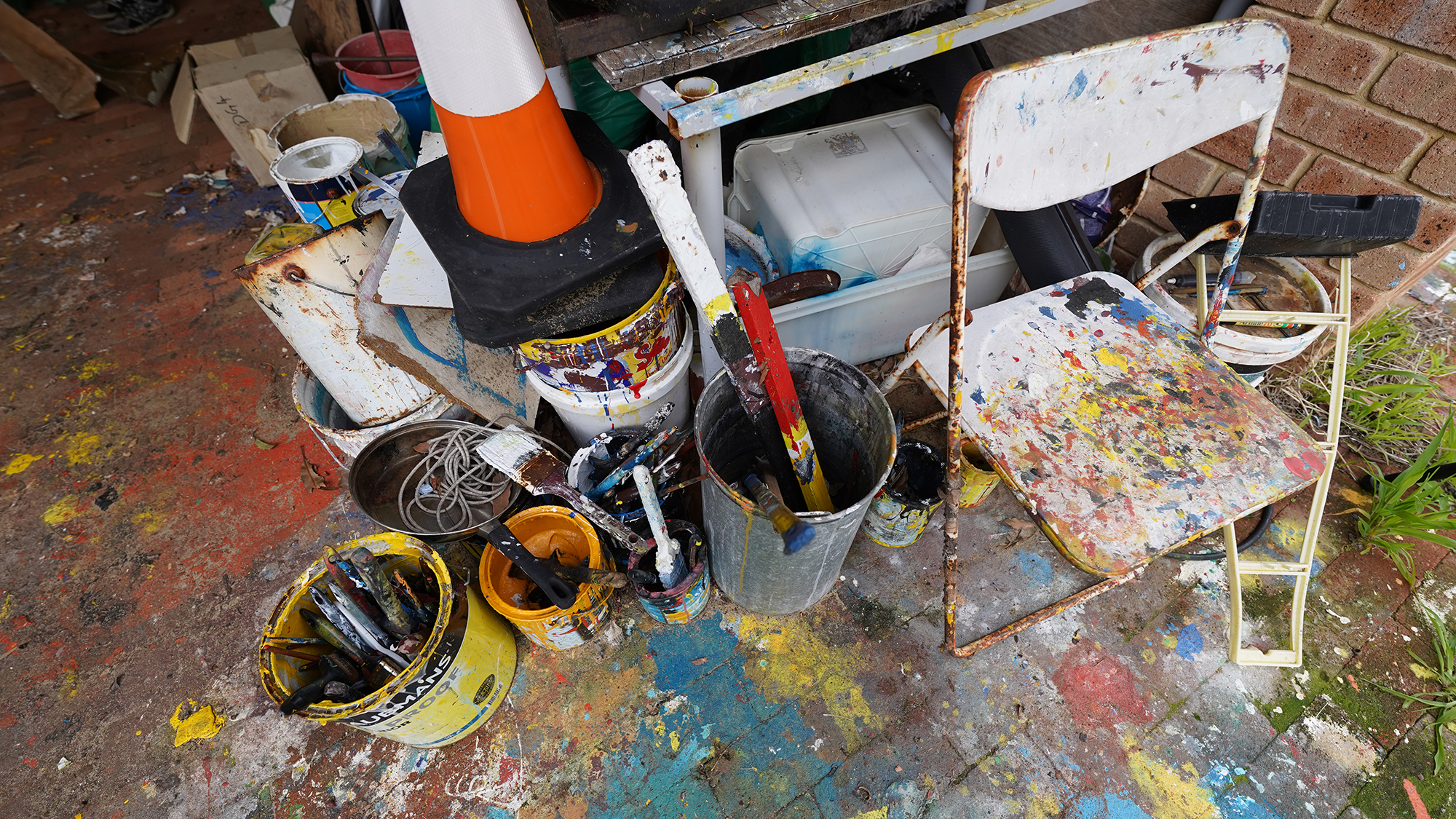 Photograph of a folding chair splattered with paint alongside buckets of paint and paintbrushes