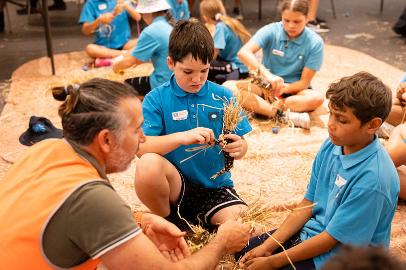 School students are sitting on the floor in groups and appear to be doing a hands on activity with straw and string. An adult sits with one group to help them.