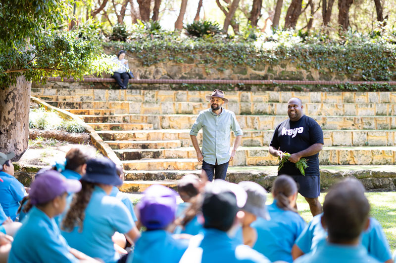 A teacher and an aboriginal man stand in front of a group of school students. they are outside in a grassy area with shady trees. they are talking to the students.