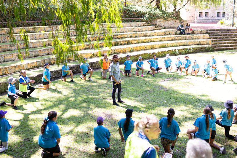 A teacher stands in the middle of a circle of students all moving in the middle of some activity. They are all in a grassy outdoor area with shady trees