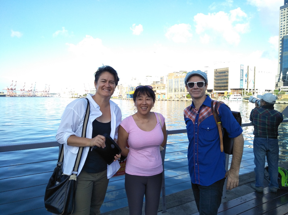 Photograph of three people standing together in front of a body of water with the outline of a cityscape behind them