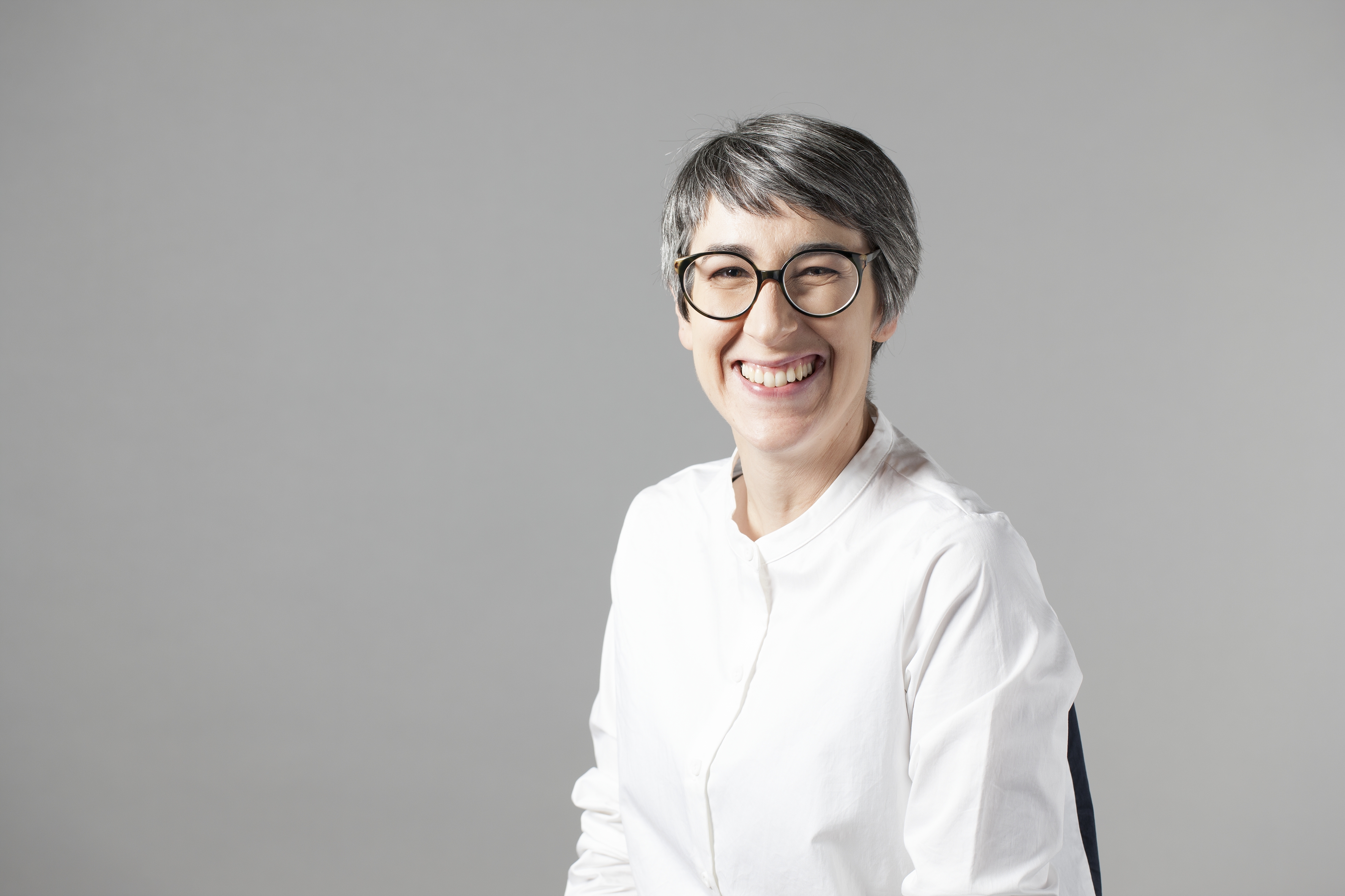 Photograph of a person seated with short grey hair and glasses and a white shirt smiling at the camera