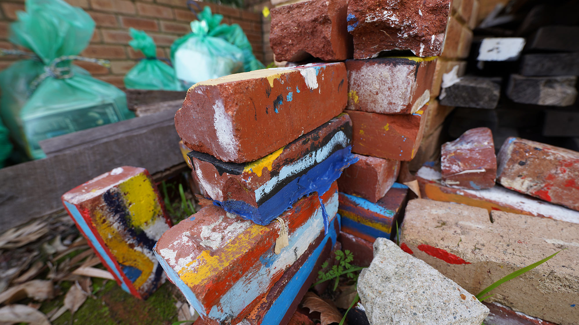 Photograph of a stack of painted bricks piled upon each other with various items, including green plastic rubbish bags, depicted in the background