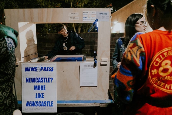 Photograph of a person within a wooden stand with a sign on it that reads 'NewsXPress Newcastle? More like Newscastle'