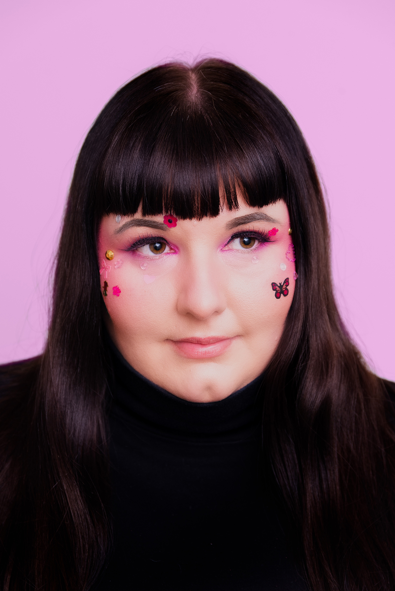 Photograph of a person with shoulder length black hair and black shirt, photographed against a pink background with stickers of butterflies, flowers and gems on their face