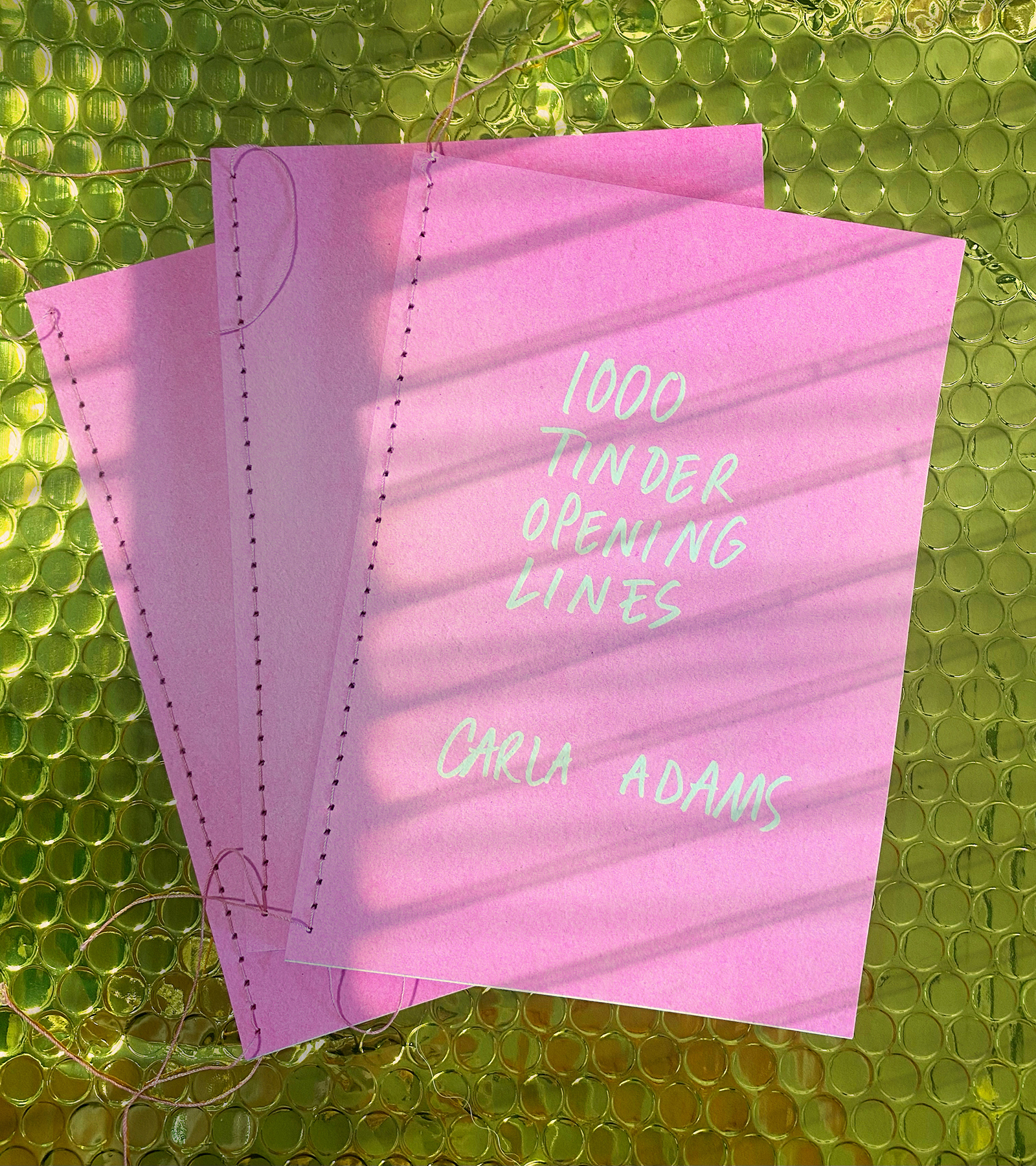 Three pink hand-sewn books titled '1000 Tinder Opening Lines, Carla Adams' on a green background
