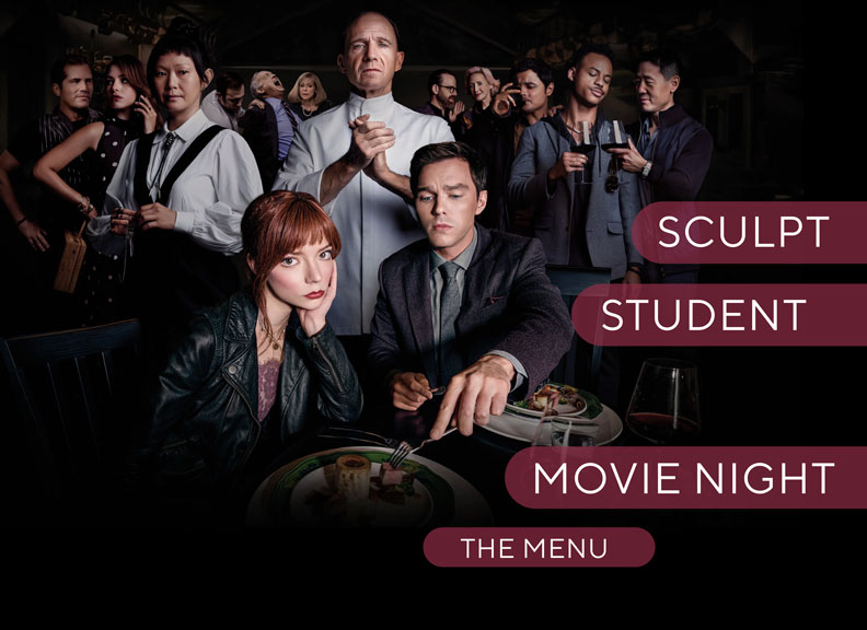A dark images shows a group of people, in the foreground there is young a couple with food in front of them, behind them stands a chef clasping his hands, other people stand behind and to the left and right of him.