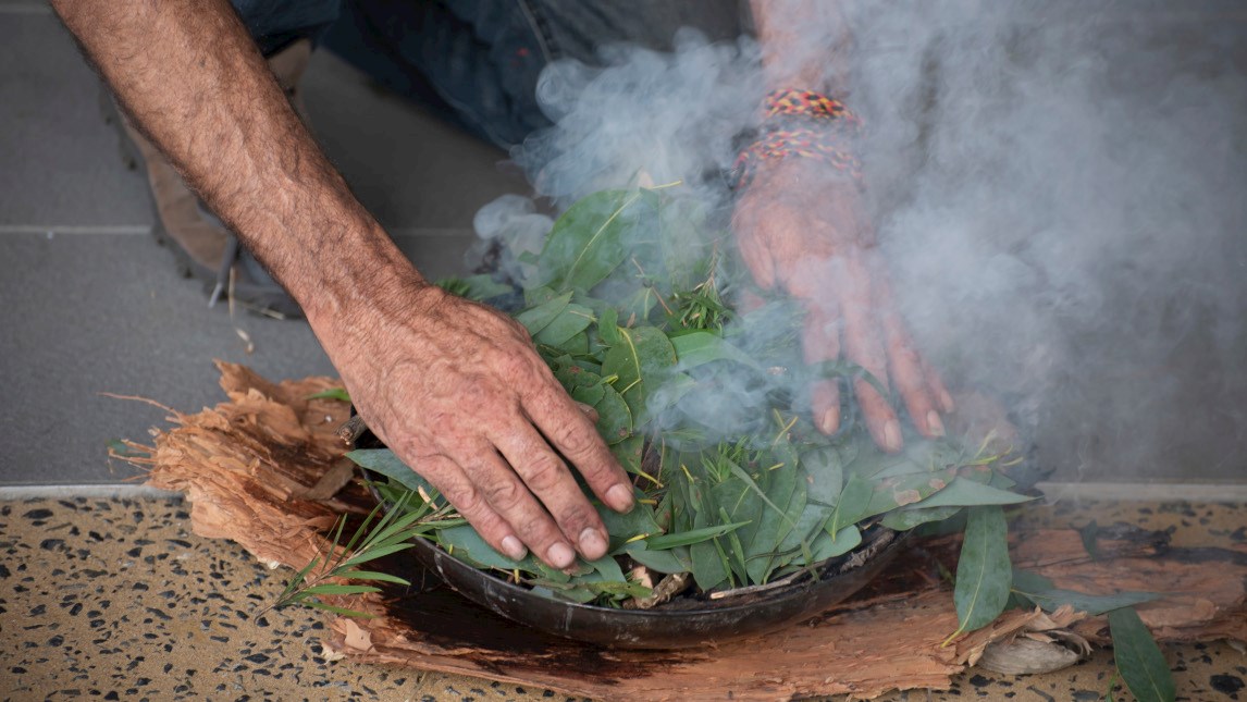 Preparation of a smoking ceremony, an Australian Aboriginal cleansing and welcome to country activity. Bark and twigs are lit in a bark container and then leaves are added to create the cleansing smoke.
