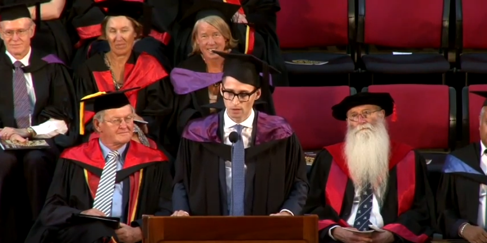 Stephen Puttick in graduation gown speaking into microphone on stage