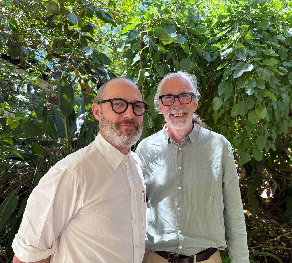 Two people standing together outdoors in a lush, sunlit garden setting.