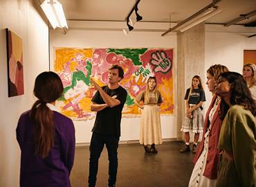 A group of visitors stands in an art gallery listening to a guide in front of a large, colourful abstract painting displayed on the wall.
