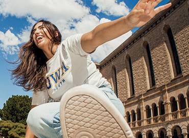 A person wearing a UWA shirt reaches toward the camera while standing outside a large stone building.