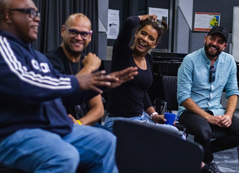 A close-up, shoulder and up image of four individuals smiling, looking at the man on the far left side. 
