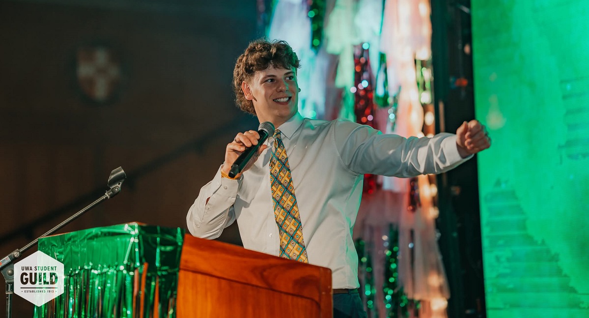 Oliver speaks into a microphone at a lectern and gestures at a screen, wearing a suit and tie, at the Guild Ball