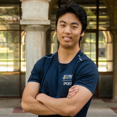 Bryan Huang stands in front of the sandstone pillars of Winthrop Hall with his arms crossed, smiling into the camera. 