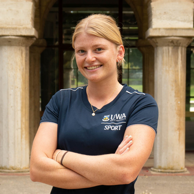 Mia Mather stands, arms folded, in front of sandstone structures at UWA