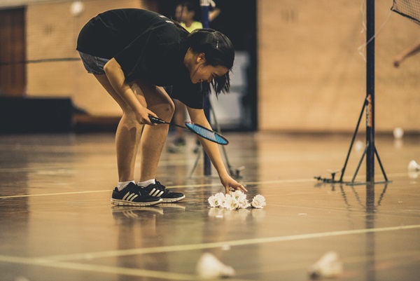 A student picks up a shuttlecock from a pile on the floor while holding a badminton racquet