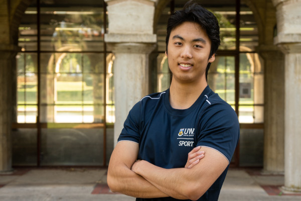 Bryan Huang stands in front of the sandstone pillars of Winthrop Hall with his arms crossed, smiling into the camera. 