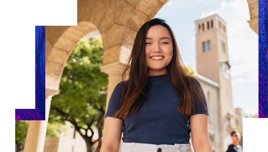 Happy UWA student stands in front of campus archway smiling at camera on a fine sunny day.
