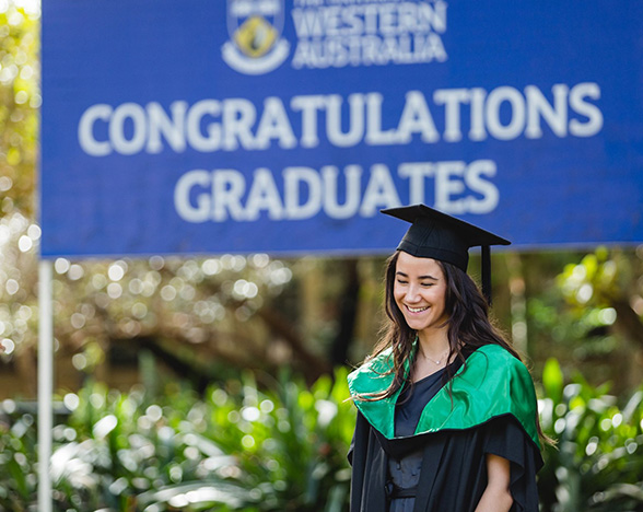 A smiling graduate outdoors in front of a sign which reads 'Congratulations graduates'