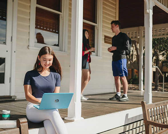 Female with laptop sitting on porch with two students talking in background