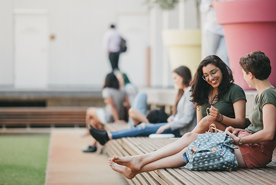 Several UWA students relax after a day of study. Two students are in the front of the image in focus the rest are blurred in the background. One student has removed their shoes and is listening to the other student tell a story.