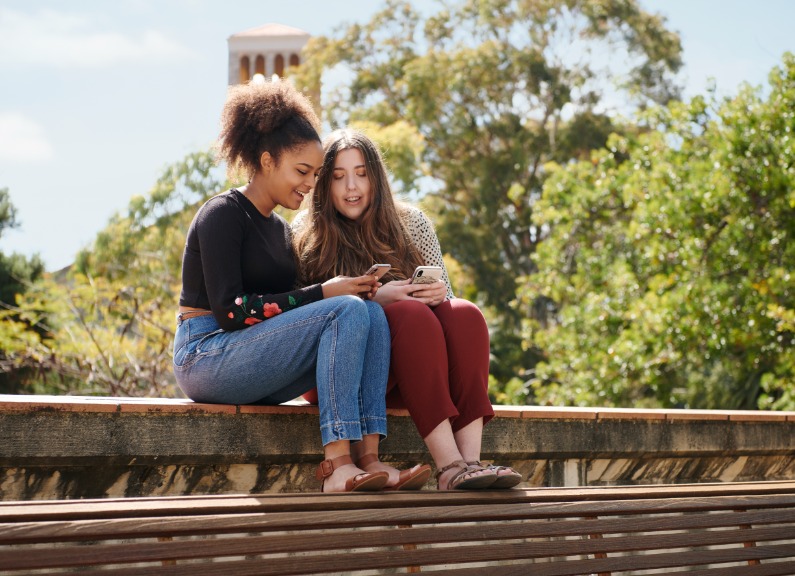 Two students sitting together looking at phone Winthrop Hall background