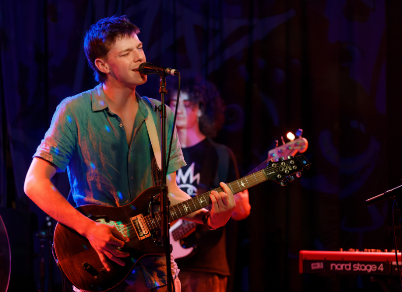 Performer singing into a microphone while playing an electric guitar on stage, with a bassist in the background and colourful concert lighting illuminating the venue.
