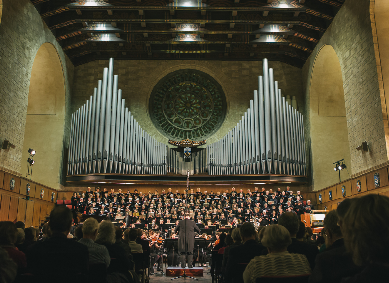 A large choir and orchestra perform before an audience beneath towering organ pipes in a grand concert hall.