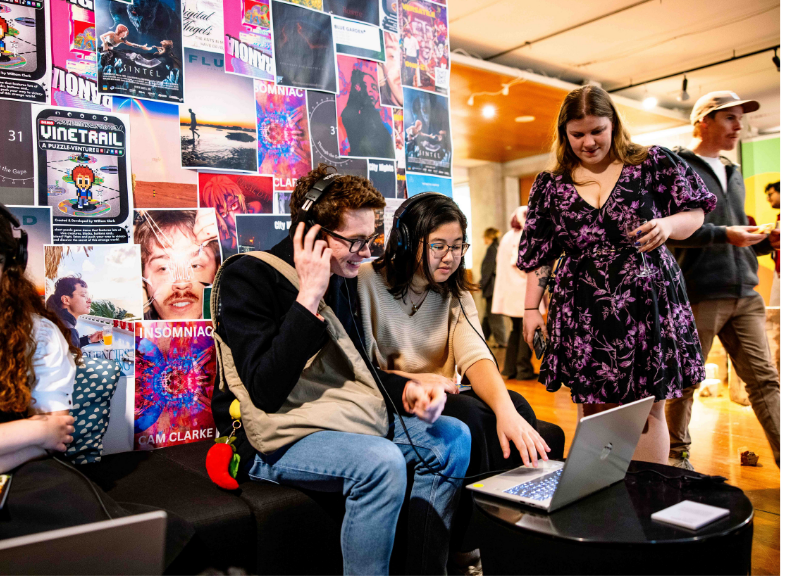 Students explore digital media projects using headphones and a laptop at a creative showcase, surrounded by colourful posters and displays.