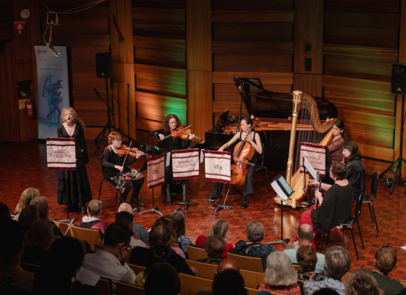 A chamber ensemble featuring strings, harp and piano performs before an audience in a concert hall, with a vocalist standing beside the musicians.