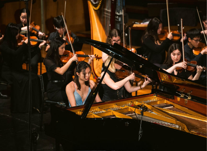 A female pianist performs at a grand piano on stage, accompanied by a chamber orchestra of string musicians during a live concert.
