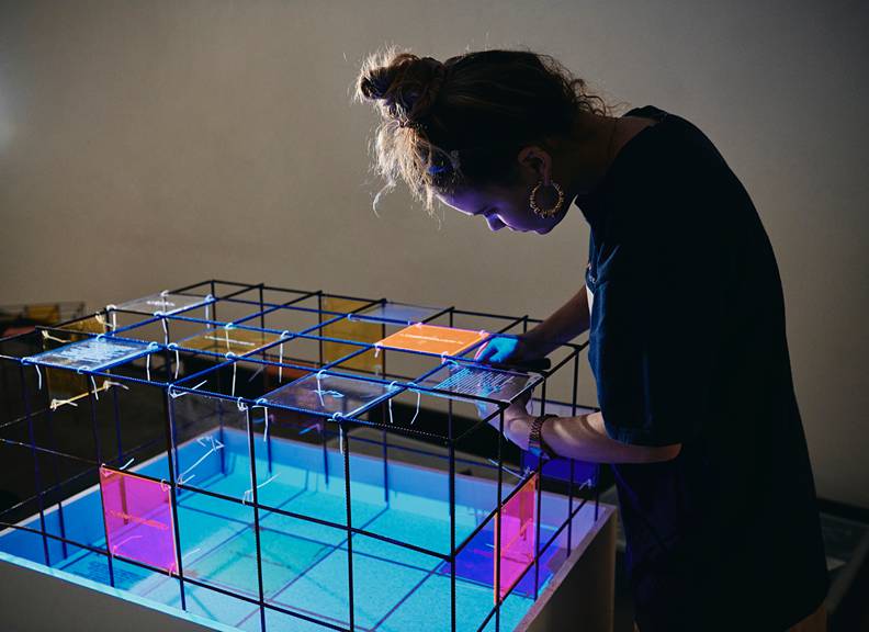 Person adjusting a colourful illuminated architectural design model made from metal grid and translucent panels in a studio exhibition setting.
