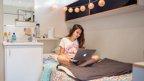 Female college resident in room sitting on bed with laptop