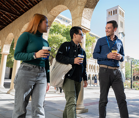 Three students with coffees walking in the archways near Hackett Hall.